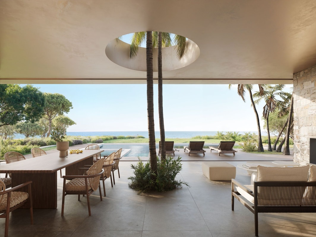 Outdoor dining and lounge area on a shaded terrace featuring a circular opening in the ceiling with a tree, overlooking a pool and the sea.
