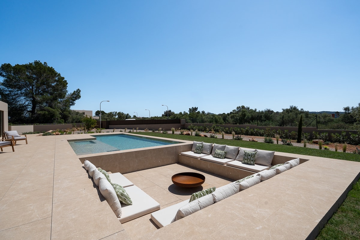 Sunken seating area on a stone terrace featuring built-in benches with cushions and a central fire pit next to a swimming pool.