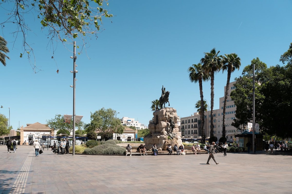Wide shot of a sunny public square in Palma with palm trees, a central equestrian monument, and pedestrians.