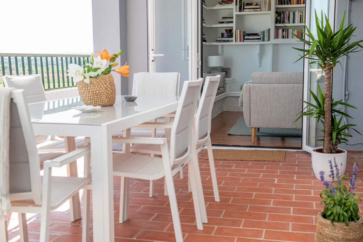 A glass-topped dining table with white chairs on a terracotta-tiled balcony, overlooking mountain views from the apartment.