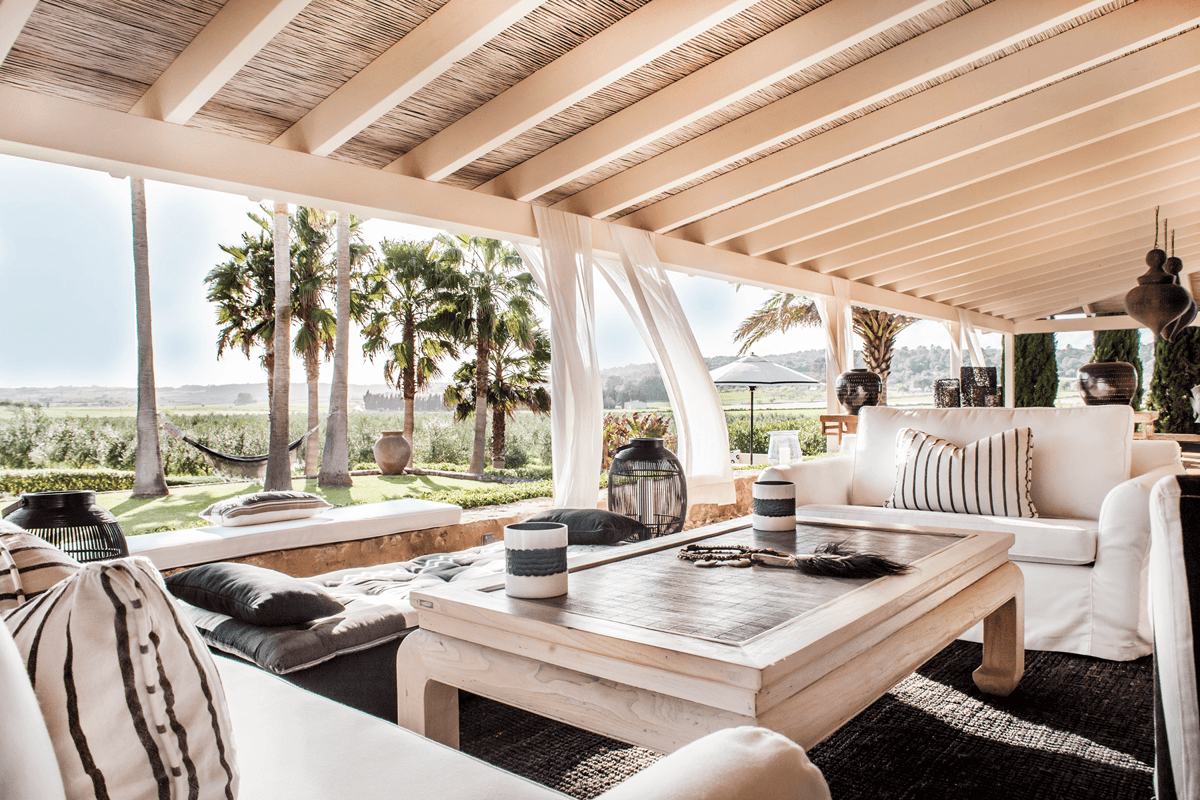 A wide, covered outdoor seating area with white fabric sofas and a wooden coffee table, overlooking a green landscape with palms.