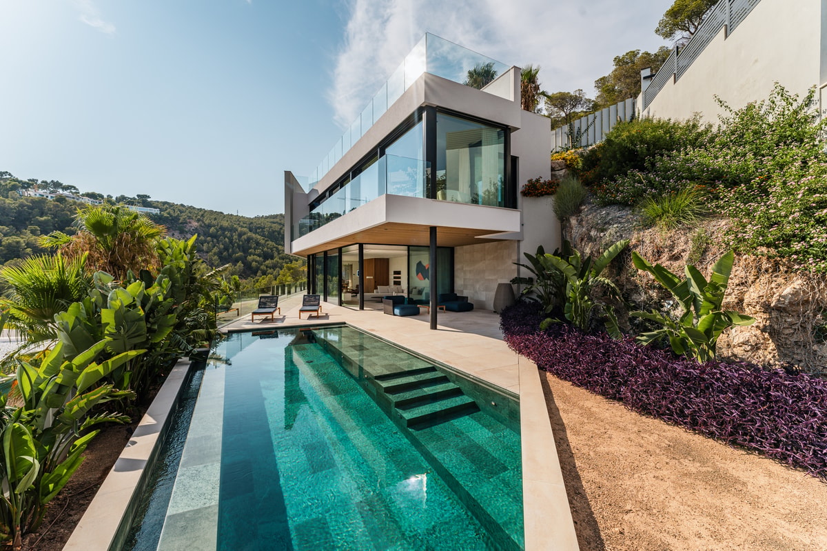 Modern two-story villa with glass walls and stone terracing, set against a rocky hill with an infinity-edge swimming pool.