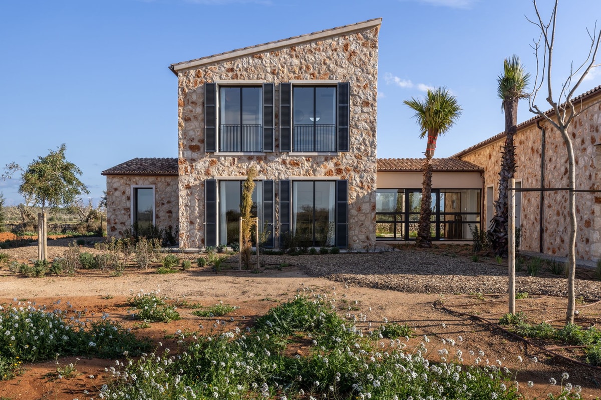 Exterior view of a two-story stone finca with a mono-pitched tile roof and dark-framed windows set in a rural landscape.