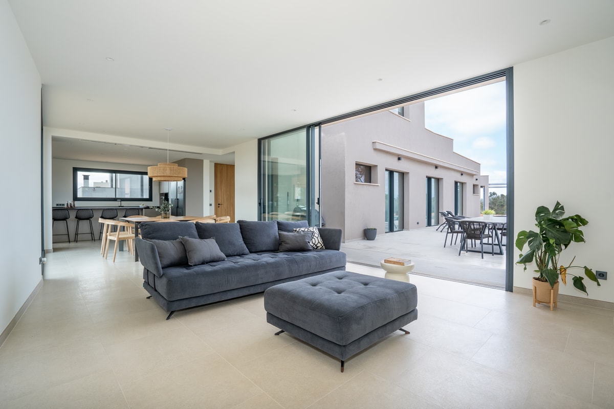 A modern living room with a dark grey sofa, light stone flooring, and sliding glass walls opening to an outdoor terrace.