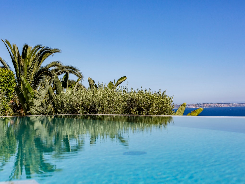 En infinity-pool med lugnt turkost vatten som reflekterar palmer och Medelhavets kustlinje.