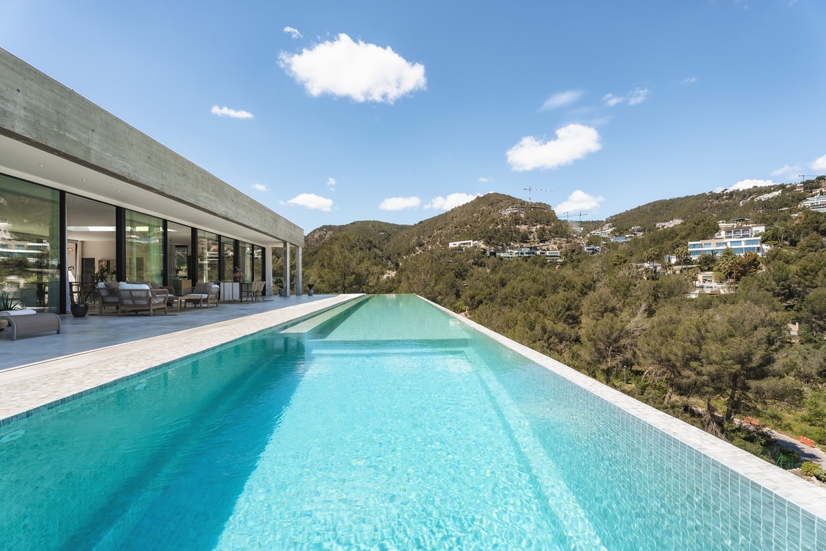 A long turquoise infinity pool runs alongside a modern villa with glass walls, overlooking a verdant mountain range.
