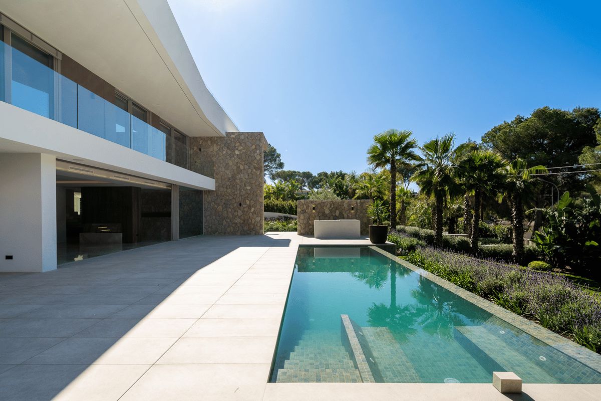 Modern villa exterior with a stone-clad wall, large terrace, and a rectangular swimming pool surrounded by palm trees.