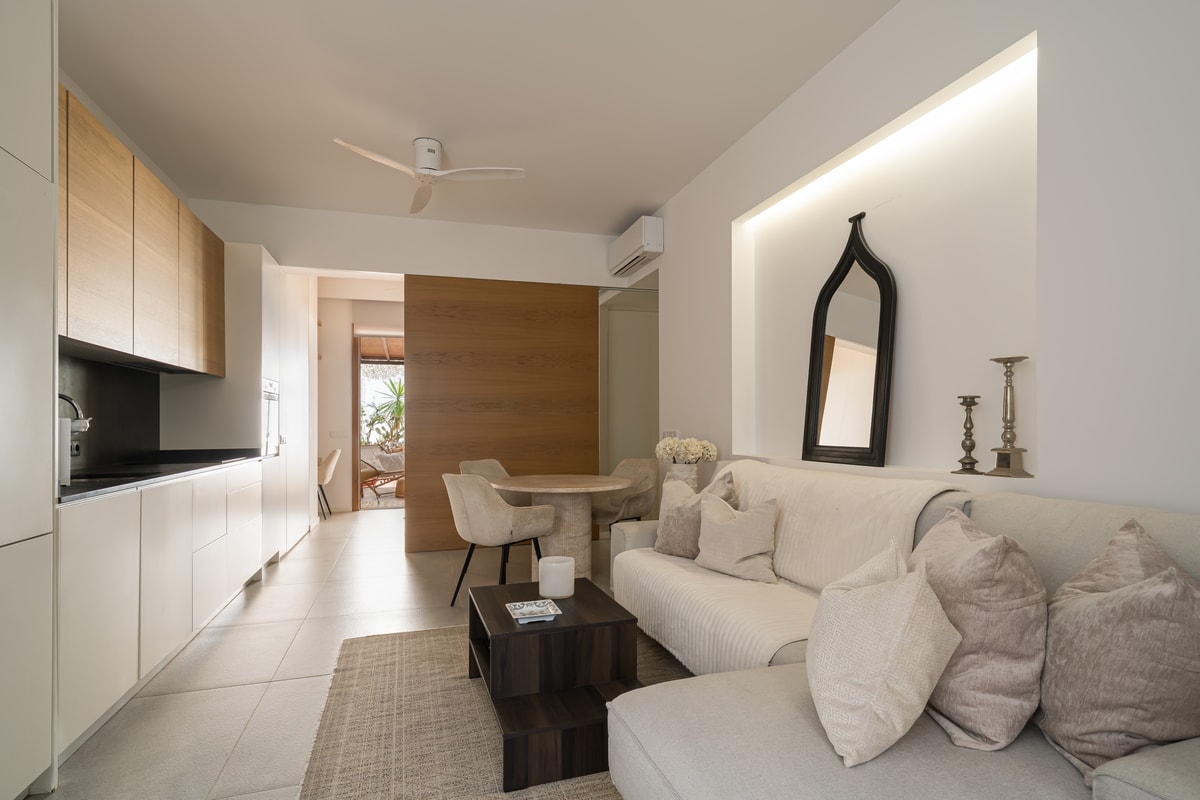 A bright apartment living space featuring a white sofa, minimalist kitchenette with wood cabinets, and dining area.