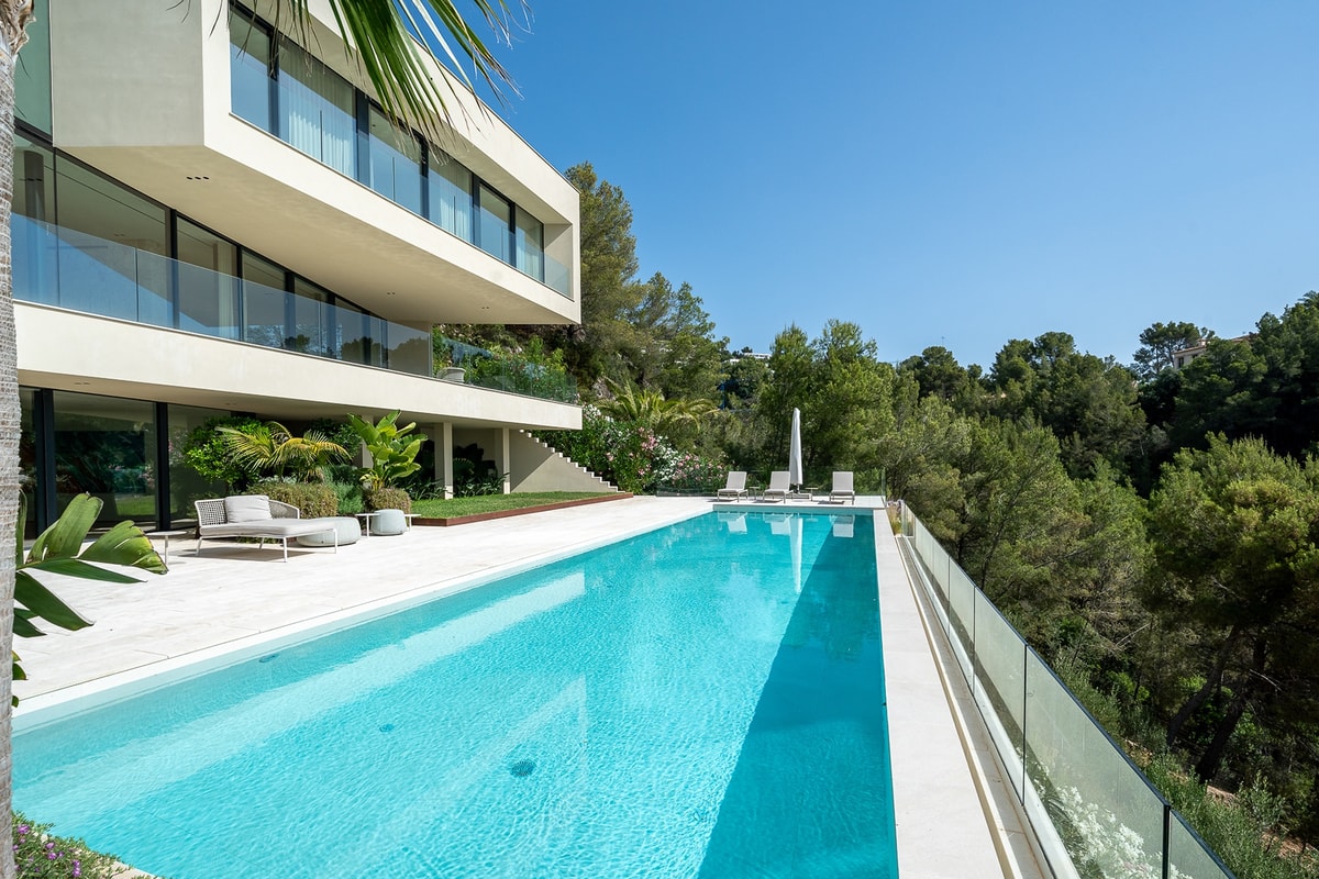 Wide exterior view of a white, multi-level modern villa featuring a long rectangular swimming pool, terrace, and trees.