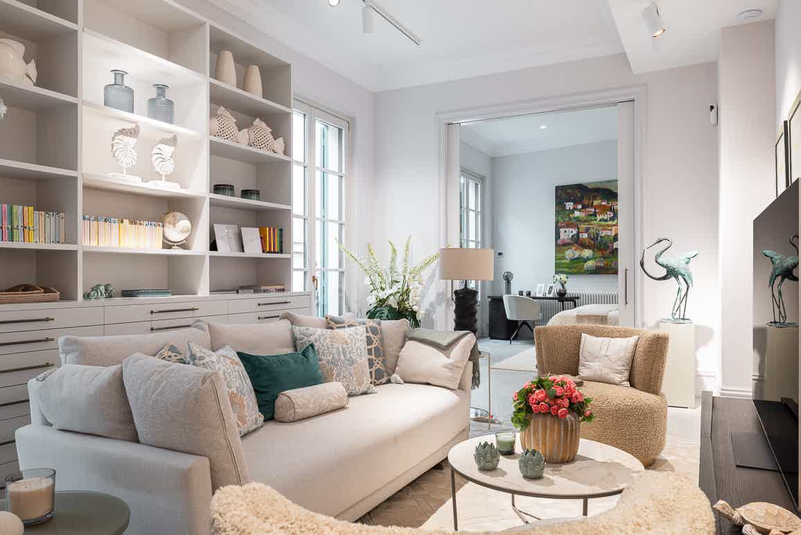 A bright living room featuring a wall-to-wall white shelving unit, a cream sofa with decorative pillows, and an open doorway leading to a secondary room.