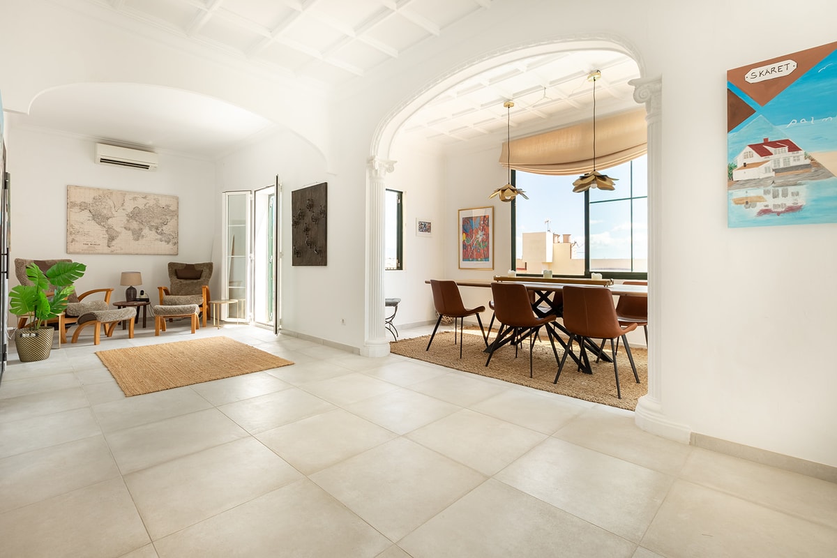 A bright living and dining room with white arched walls, tiled floors, a wooden dining set, and a cozy seating area.