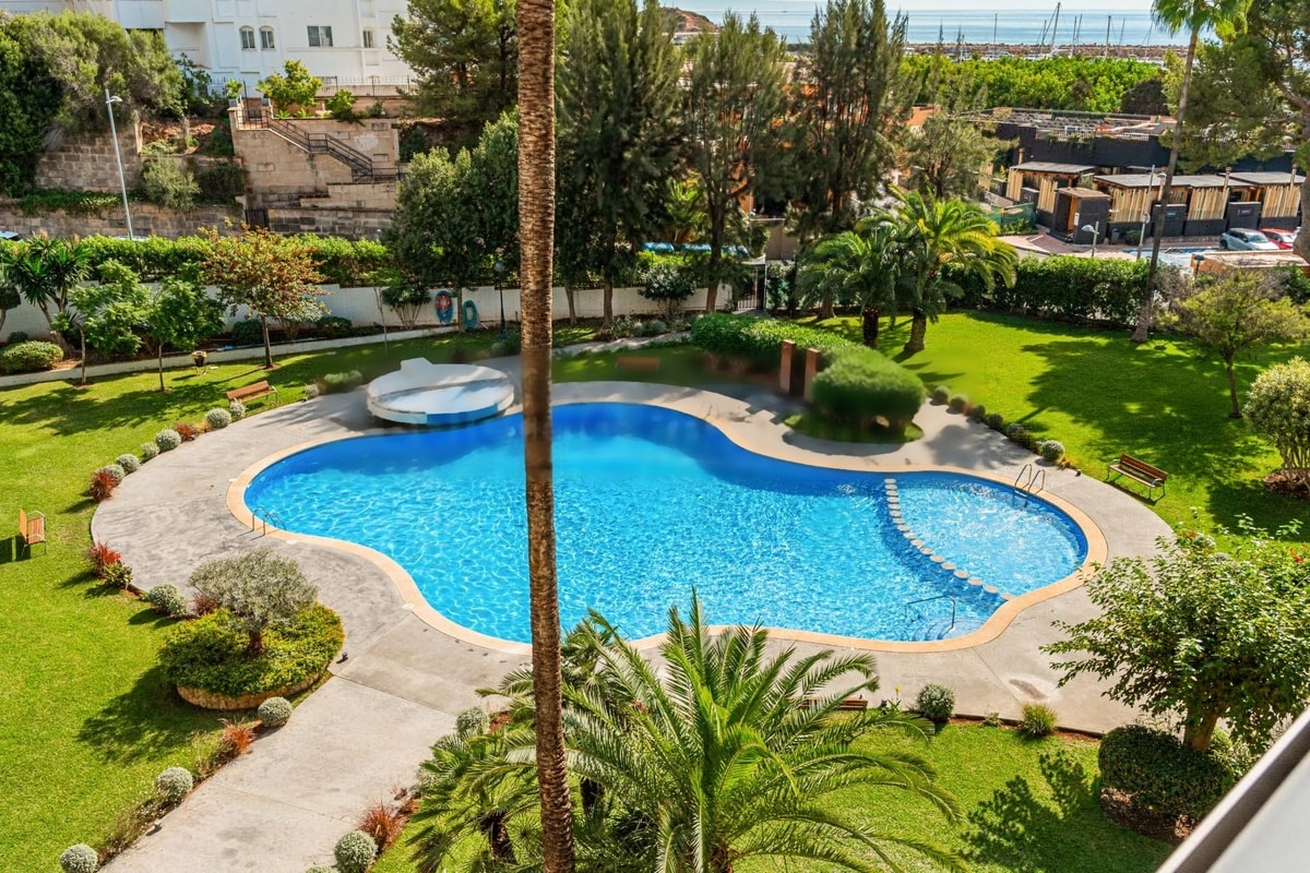 High-angle view of a kidney-shaped blue swimming pool surrounded by manicured green lawns and palm trees in Portals Nous.