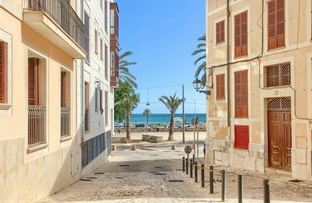A narrow cobblestone street in a historic Old Town, framed by pale-colored residential buildings with wooden shutters, leading directly to a blue sea view.