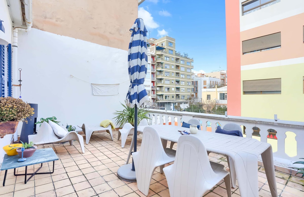 A sunny outdoor terrace with a dining table, chairs, and a blue-striped parasol, featuring city views of neighboring buildings.