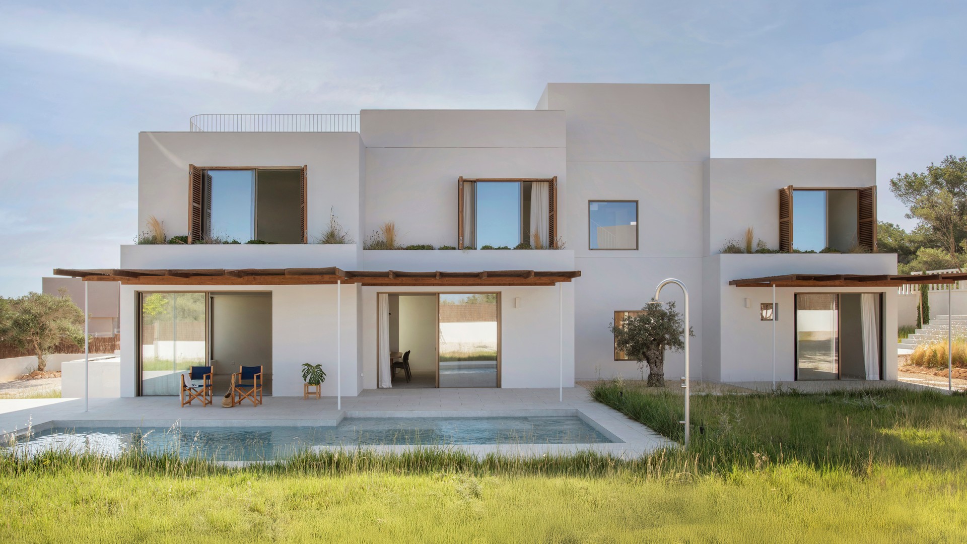 Modern white two-story villa with wooden shutters and a rectangular pool reflecting the sky, bordered by green grass.