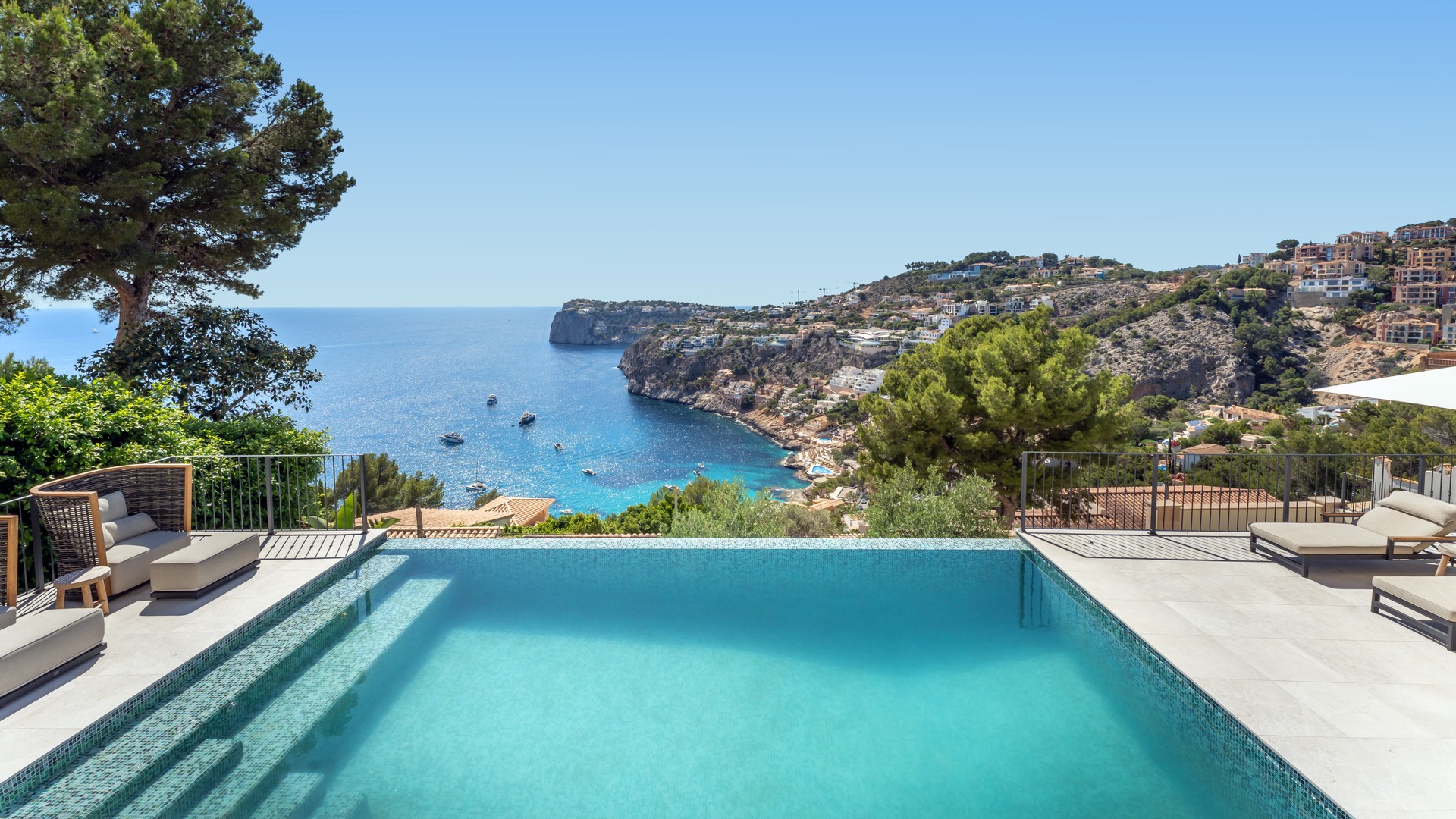 An infinity edge swimming pool with turquoise mosaic tiles overlooking a sea bay with rocky cliffs and pine trees.