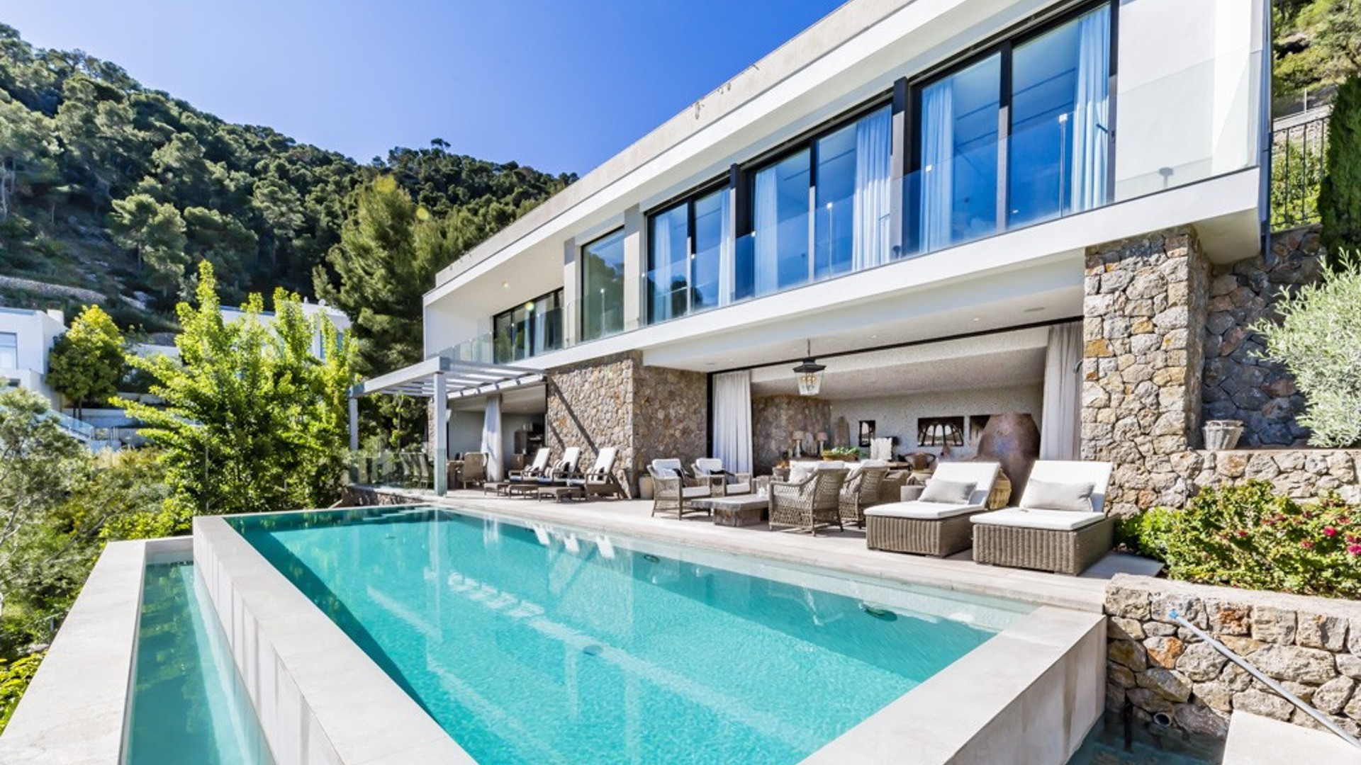 Exteriör av en modern vit villa med stenfasad, infinity-pool med turkost vatten och en möblerad terrass.