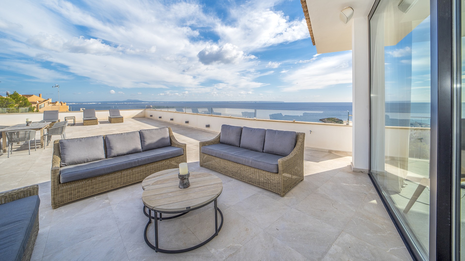 A wide outdoor terrace featuring woven lounge furniture, stone flooring, and expansive views of the sea under a blue sky.
