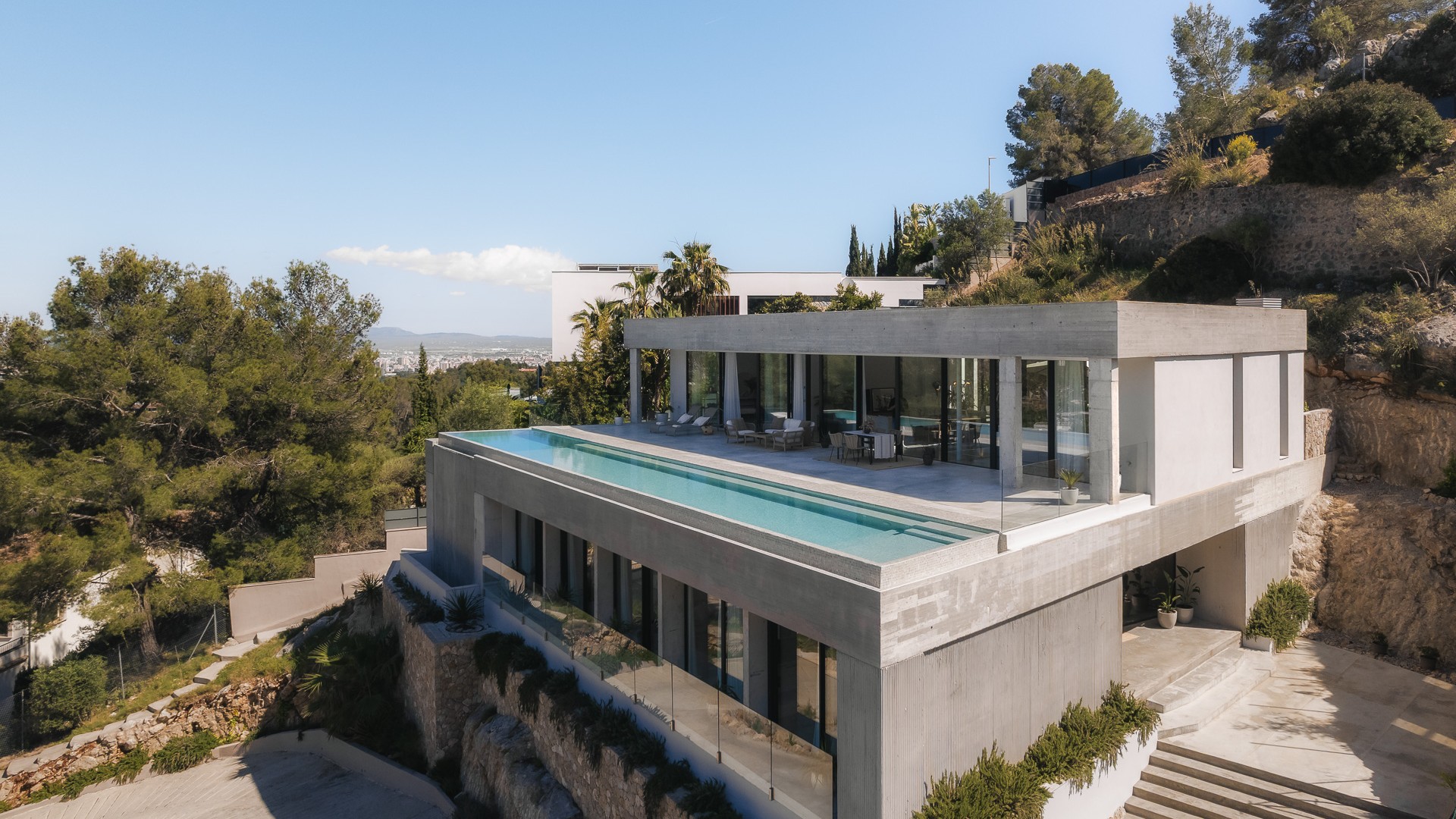 Elevated view of a contemporary concrete villa featuring a rooftop infinity pool and floor-to-ceiling glass walls.