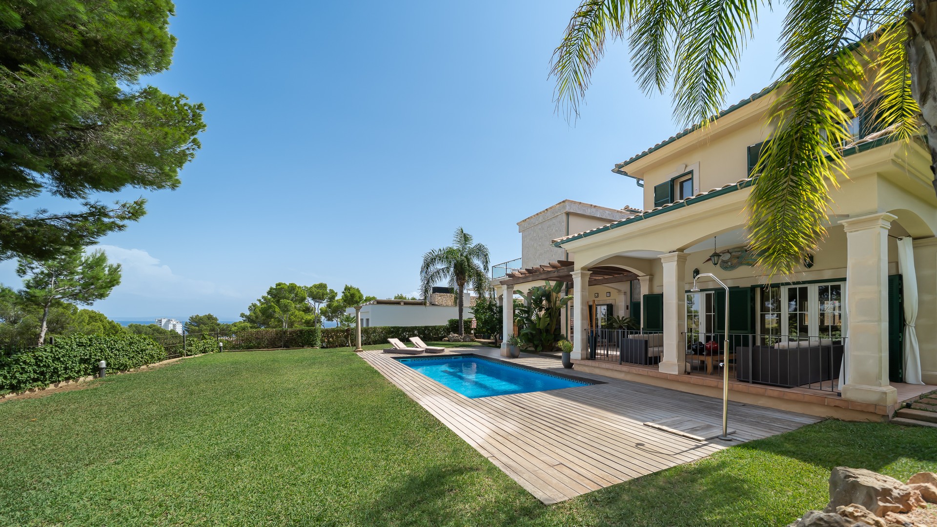 Exterior view of a villa with a rectangular swimming pool, manicured lawn, sandstone columns, and a shaded terrace.