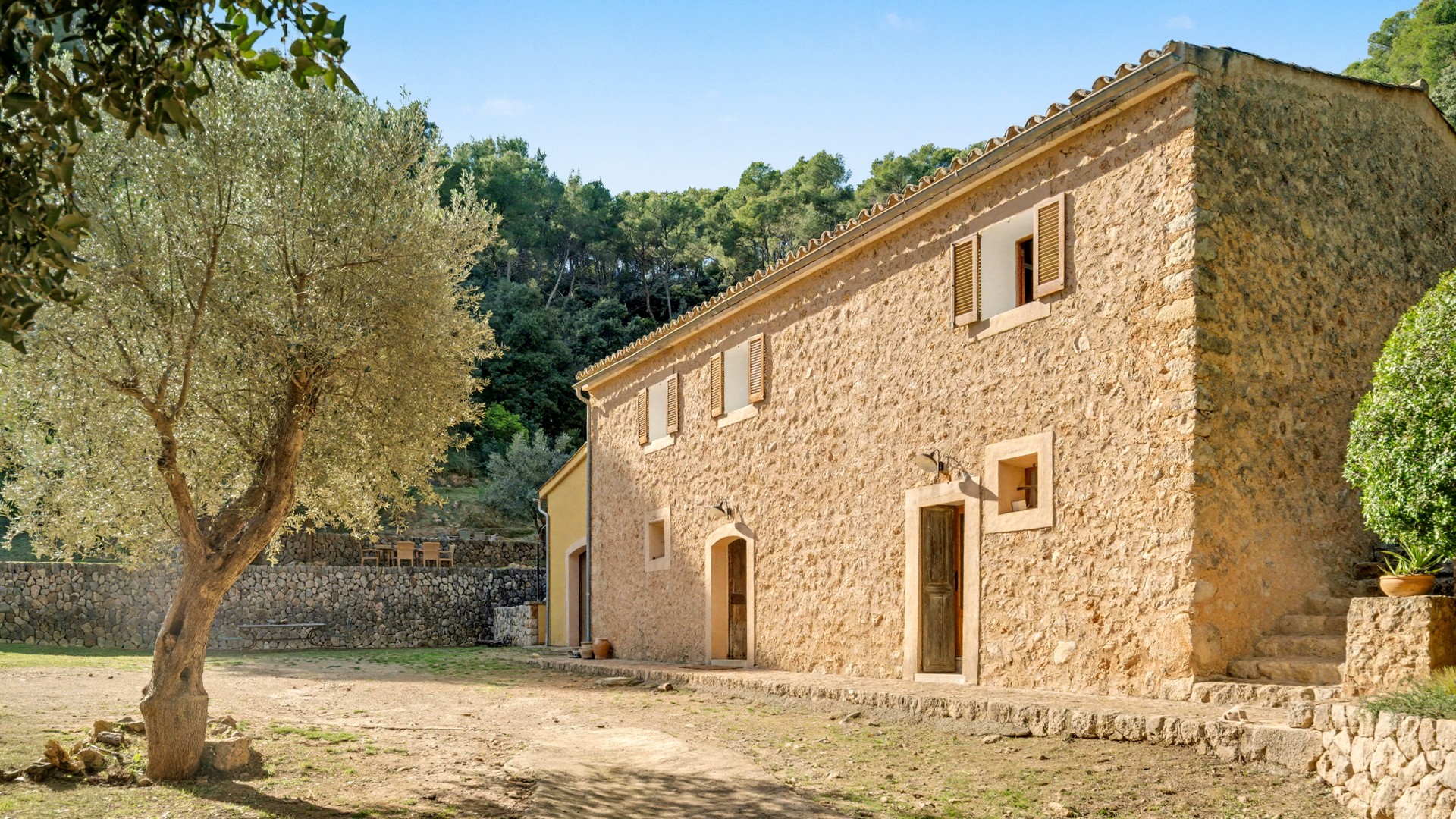 Exterior view of a traditional stone finca with timber shutters, surrounded by olive trees and pine forest under a blue sky.
