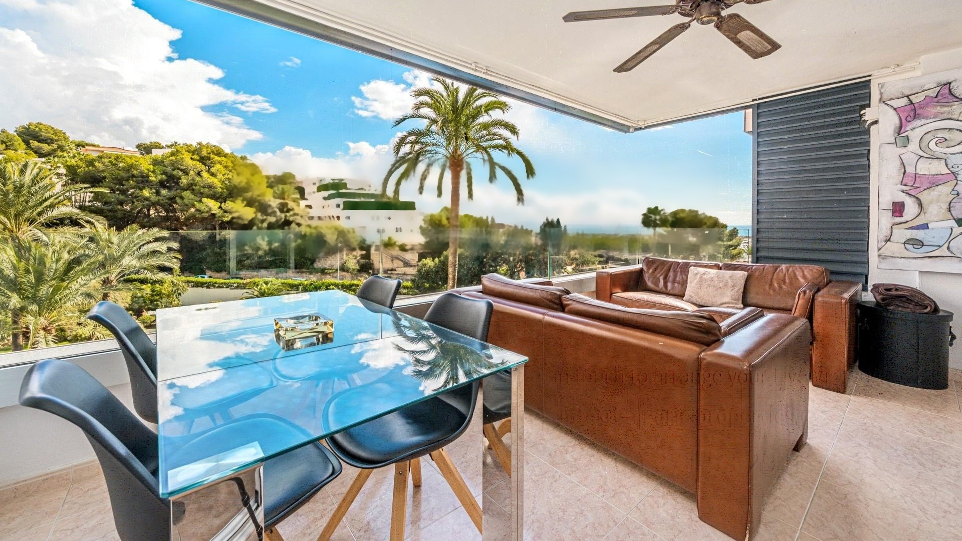 A covered terrace with a glass dining table, leather sofa, and ceiling fan overlooking a coastal landscape with palm trees.
