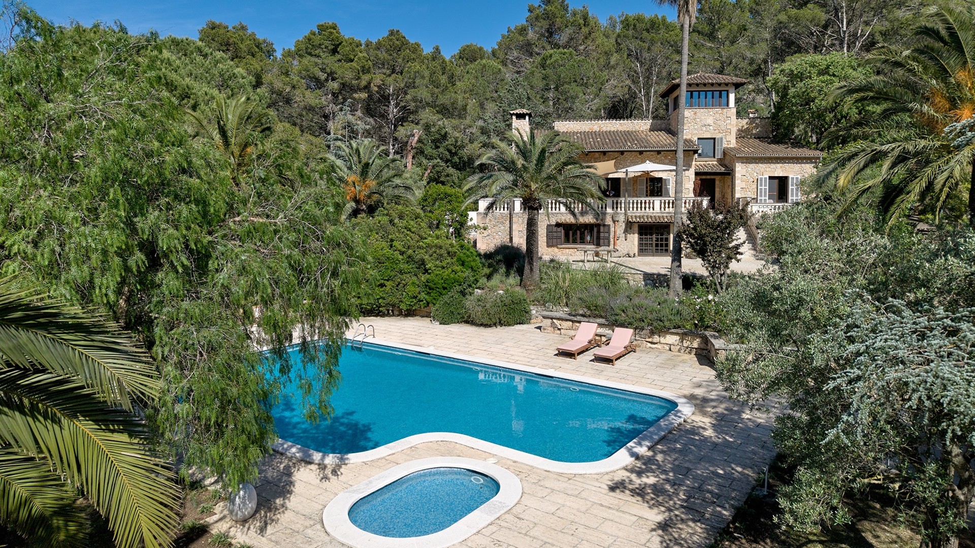 Exterior view of a traditional stone finca surrounded by trees, featuring a rectangular swimming pool and a separate plunge pool on a stone patio.