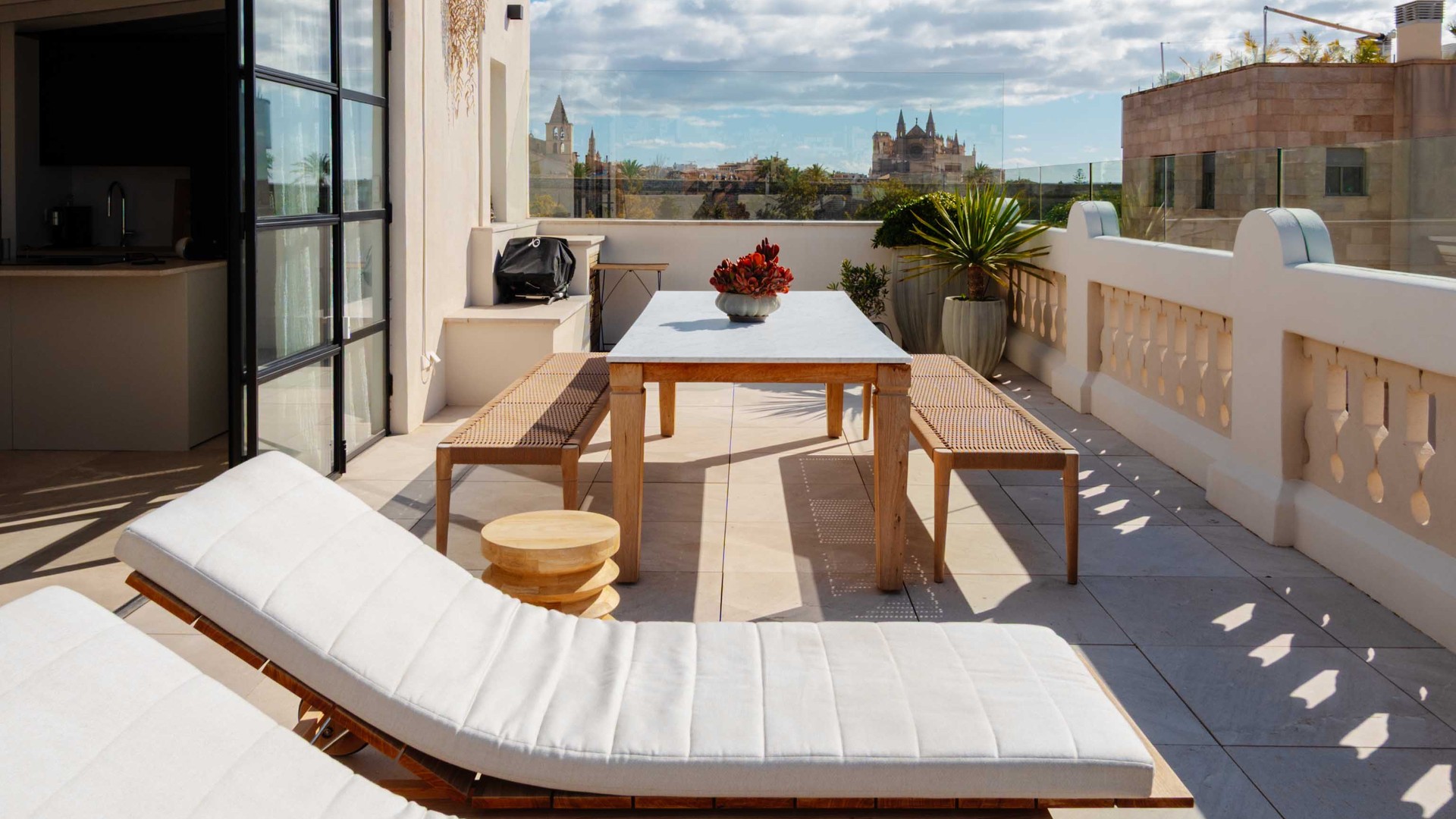 An expansive outdoor rooftop terrace featuring white sun loungers, a wooden dining table, and a view over the Santa Catalina skyline under a clear blue sky.