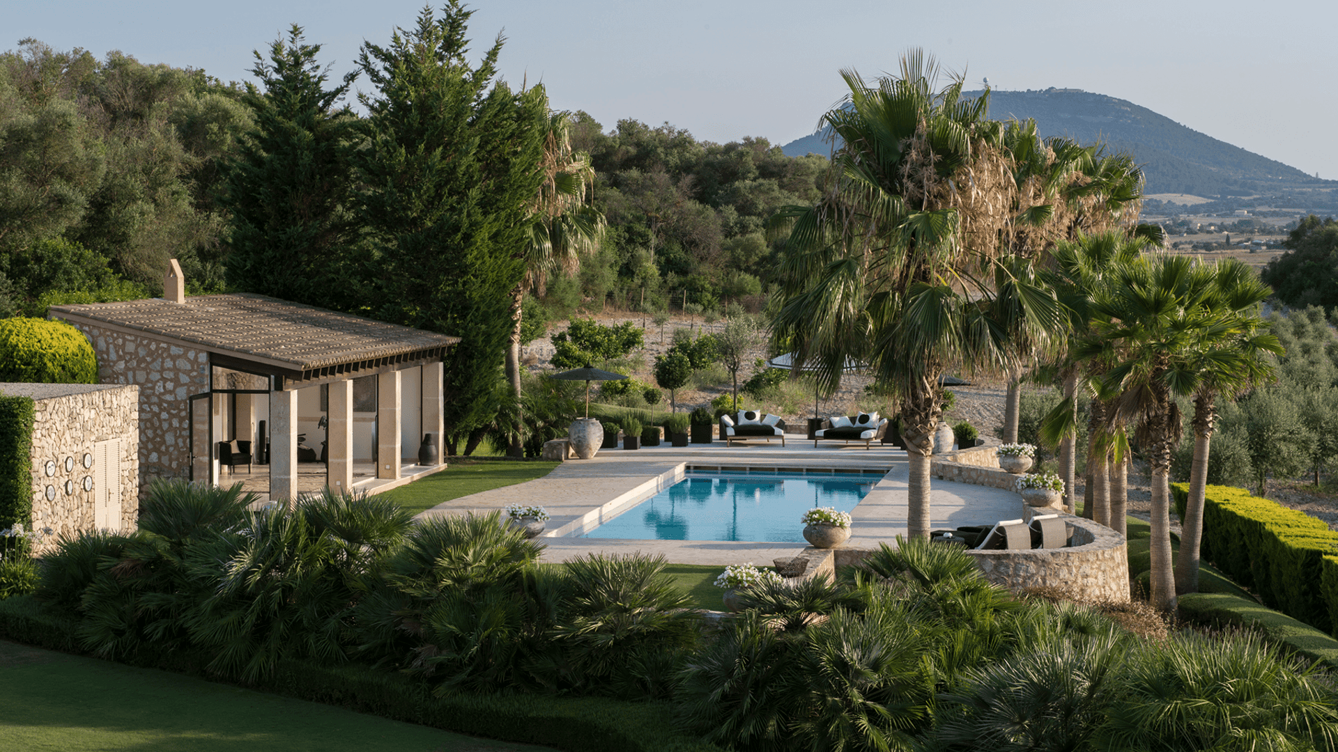 A stone-built finca exterior featuring a rectangular swimming pool, landscaped garden, and mountain views in Algaida.