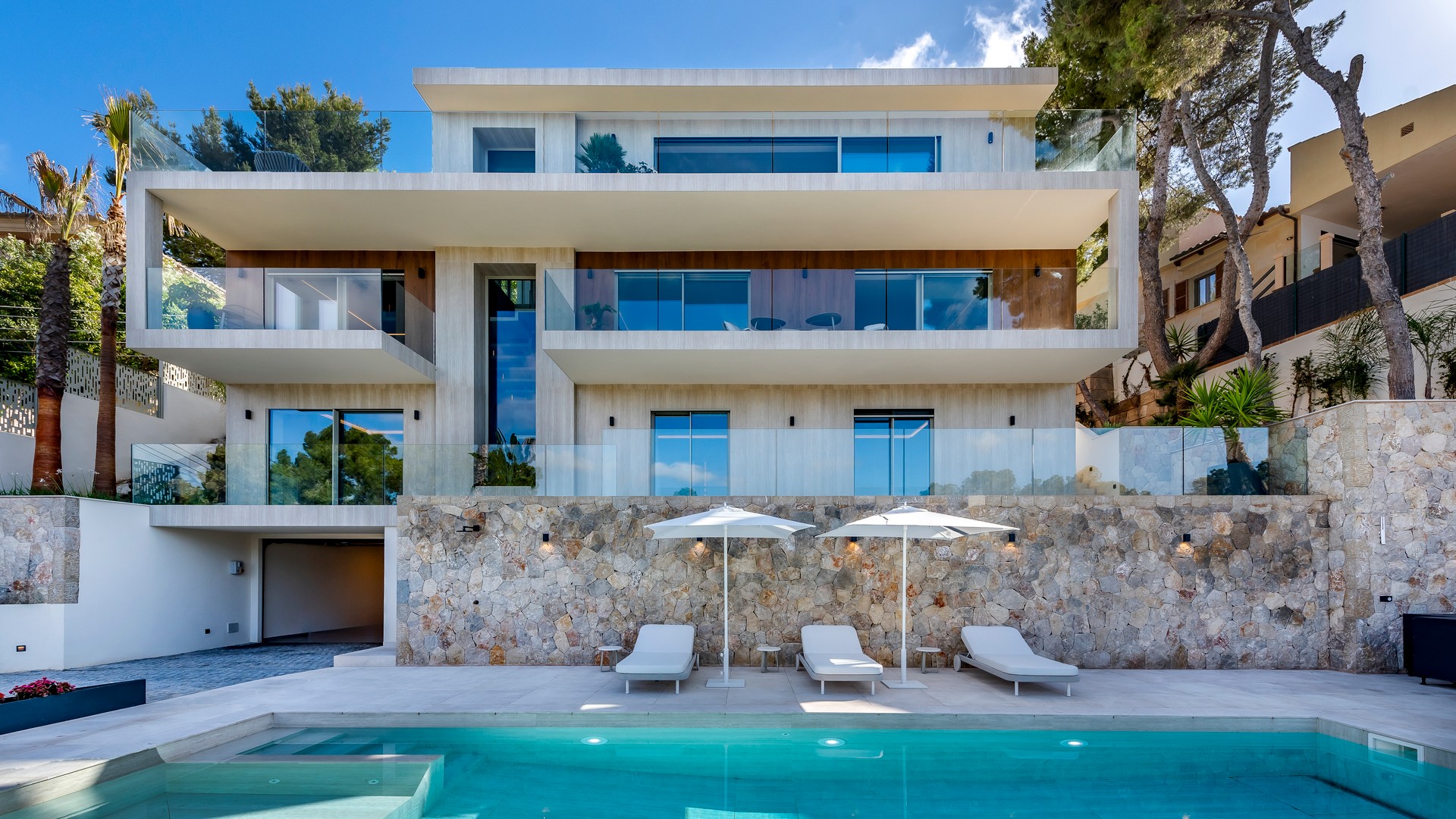 Three-story modern villa with glass railings, stone retaining wall, swimming pool, and sun loungers on a paved terrace.