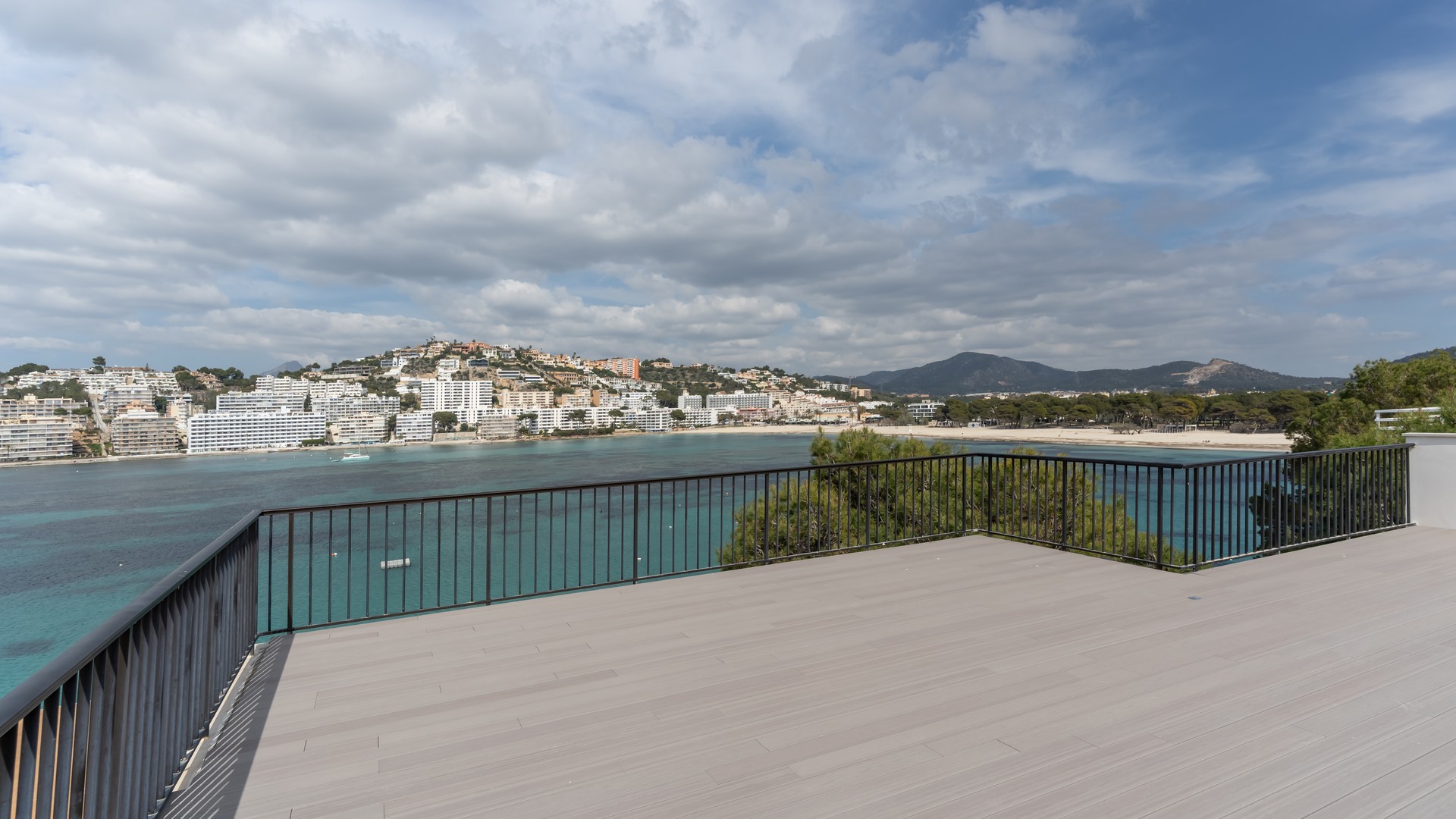 A wide grey-decked terrace with black metal railings overlooking a Mediterranean coastal town, sandy beach, and mountains.