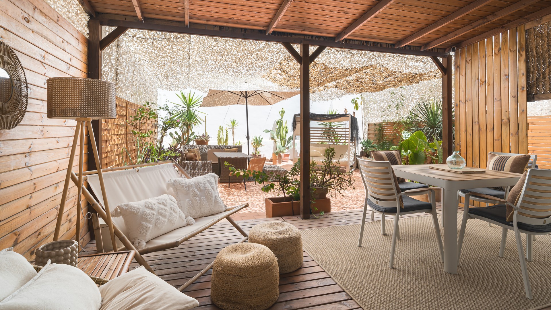 A shaded outdoor terrace with wooden panel walls, dining furniture, a sofa, and potted plants under a perforated roof.