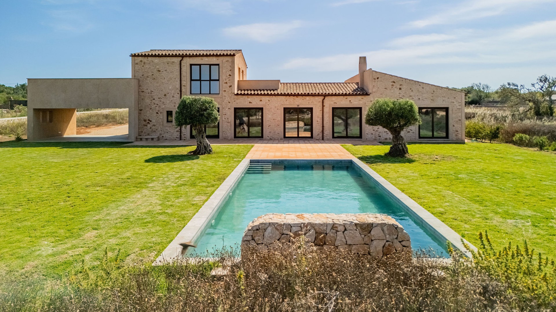 Exterior view of a stone finca in Santanyí featuring a rectangular swimming pool, manicured lawn, and large glass doors.