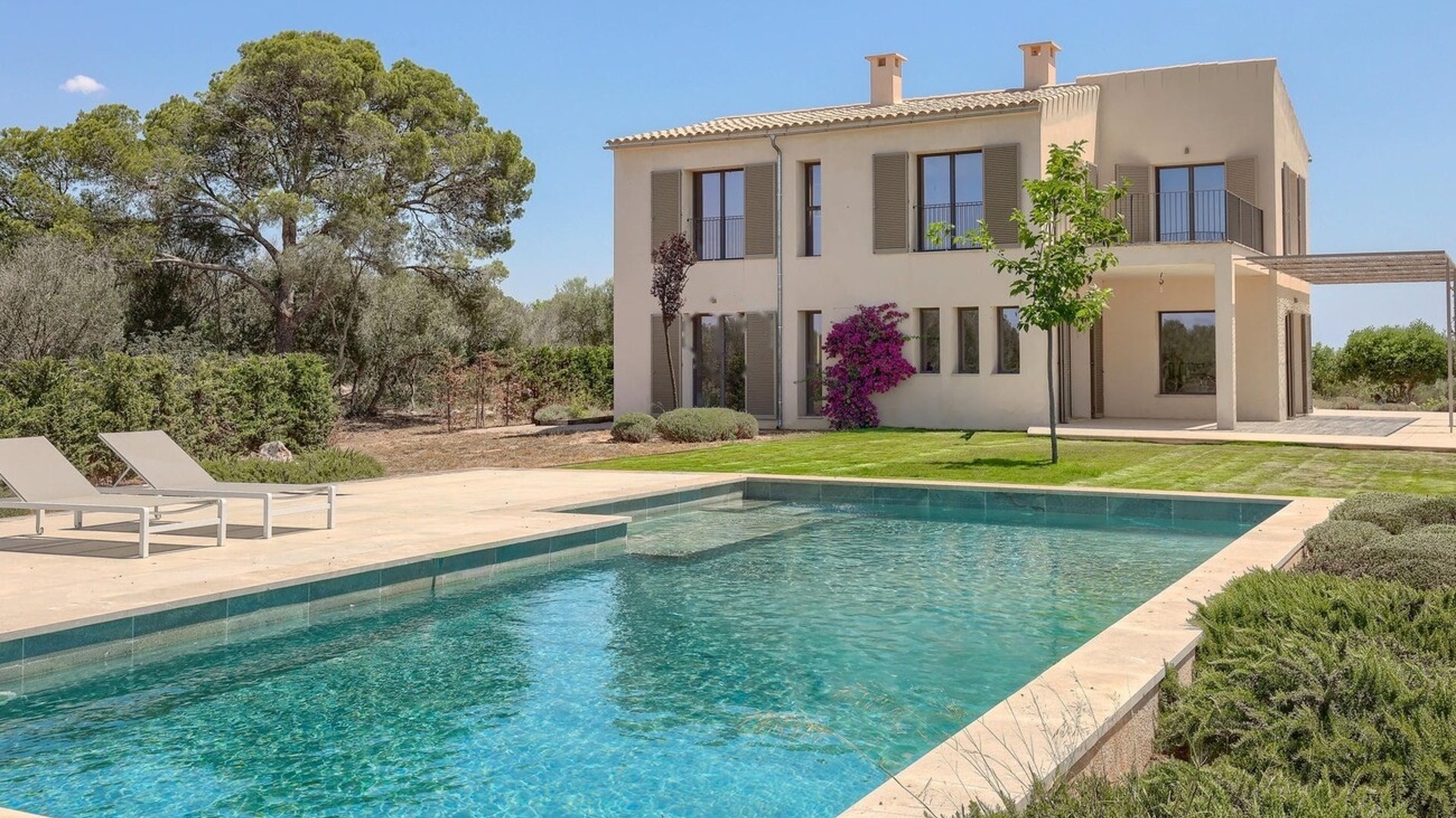 Exterior view of a beige stone finca with wooden shutters, a swimming pool, and lounge chairs in a landscaped garden.