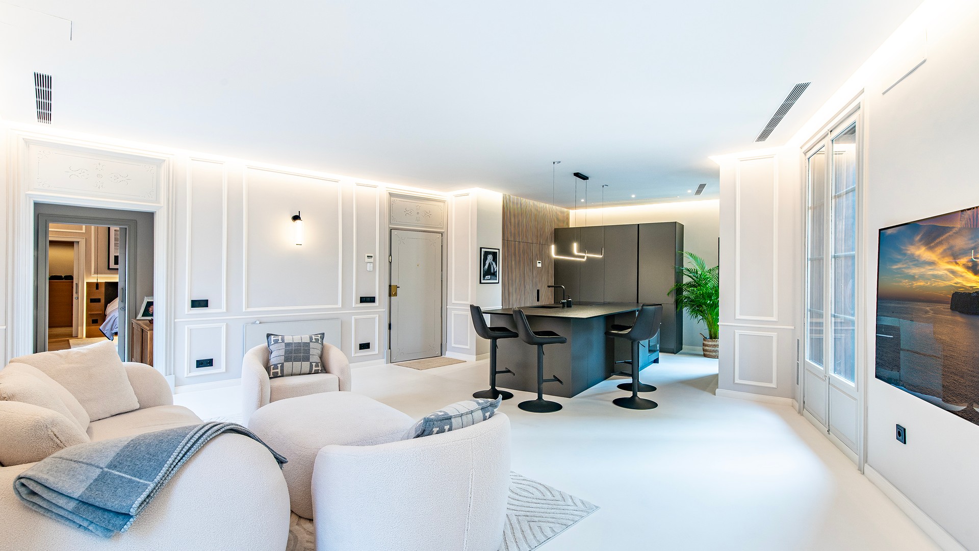 Wide shot of a modern white open-plan living room with a kitchen island, textured armchairs, and floor-to-ceiling windows.