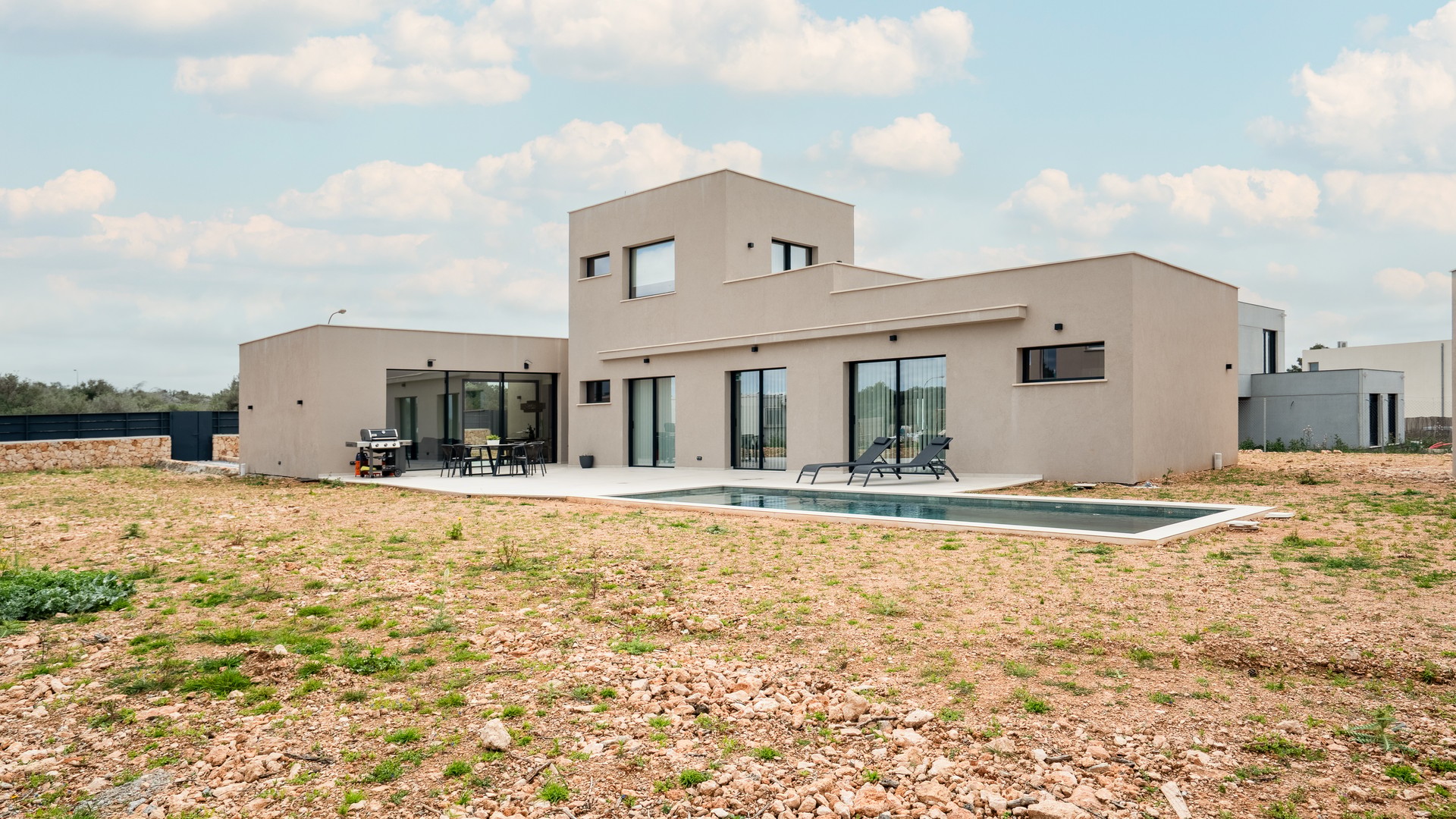Exterior view of a modern two-story beige villa with a swimming pool, flat roof, and large glass sliding doors.