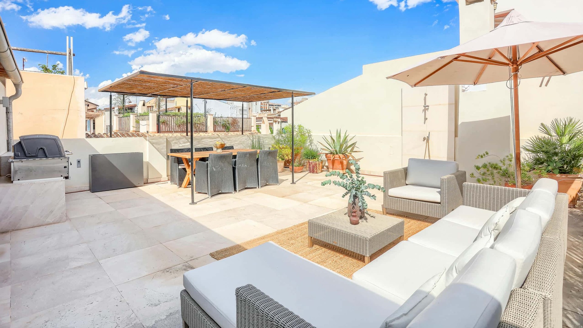 A bright rooftop terrace with a dining area under a wooden pergola and a lounge seating area with white cushions and a large cream umbrella under a clear blue sky.