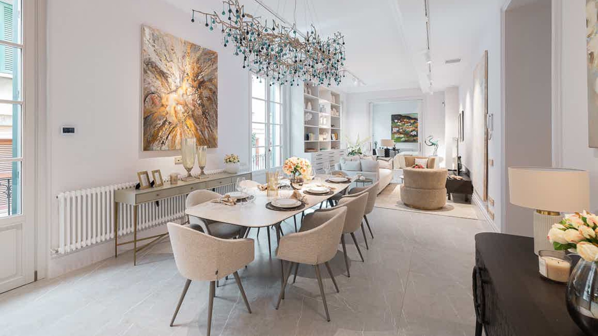 Dining area with a rectangular light-wood table, cream upholstered chairs, and a large sculptural chandelier overhead.