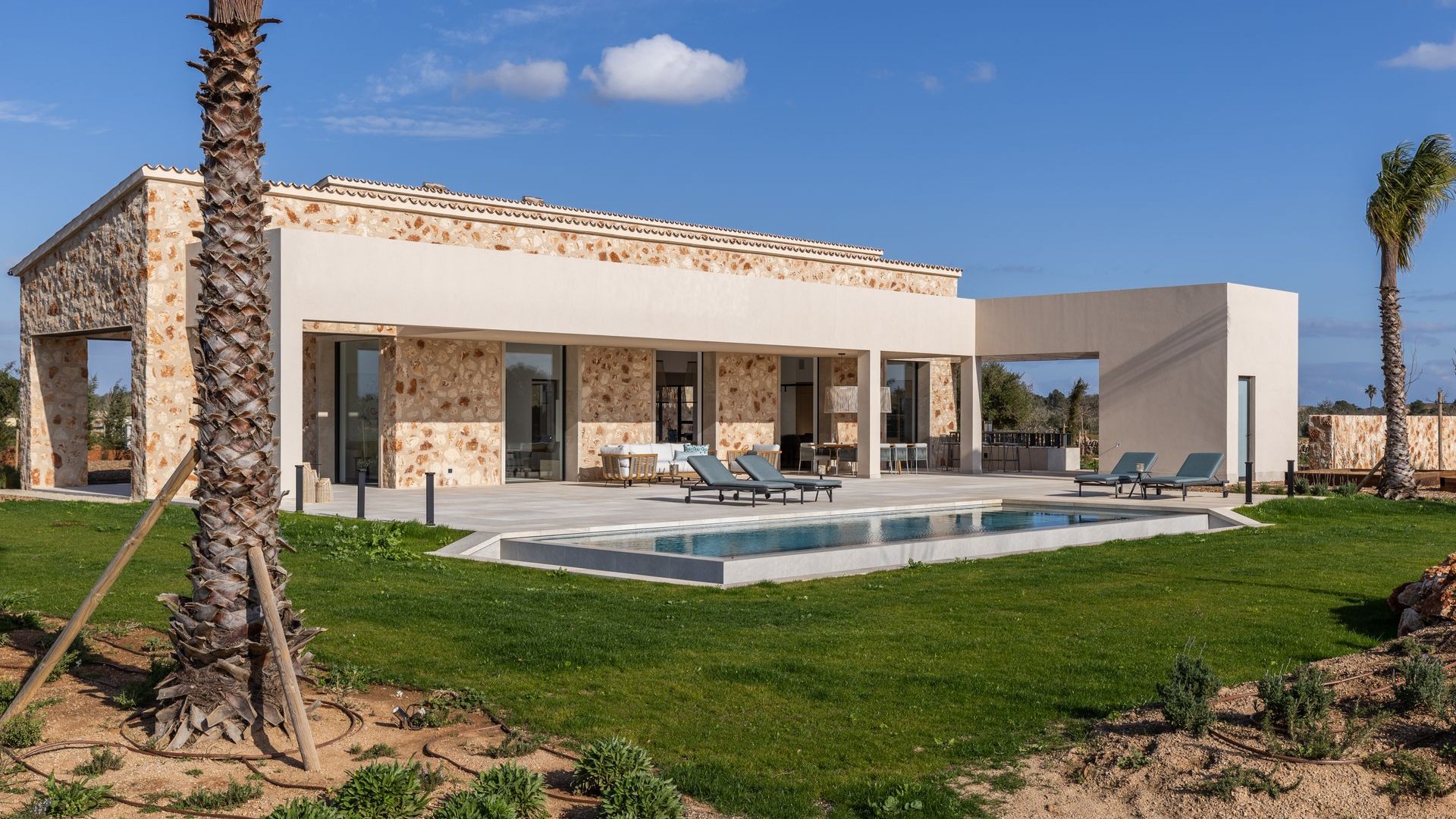 Exterior view of a single-story stone finca in Santanyí with a swimming pool, lawn, and pergola-covered terrace.