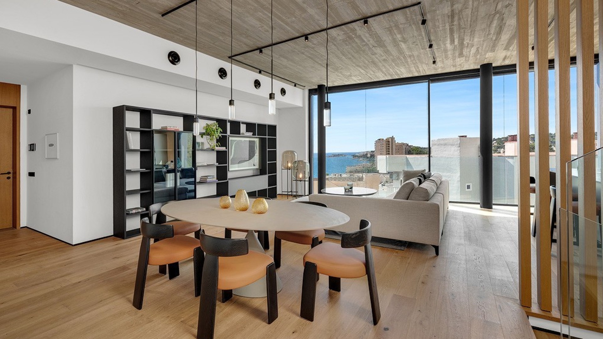 Modern open-plan living and dining room with concrete ceiling, wooden floors, and floor-to-ceiling windows overlooking the sea.