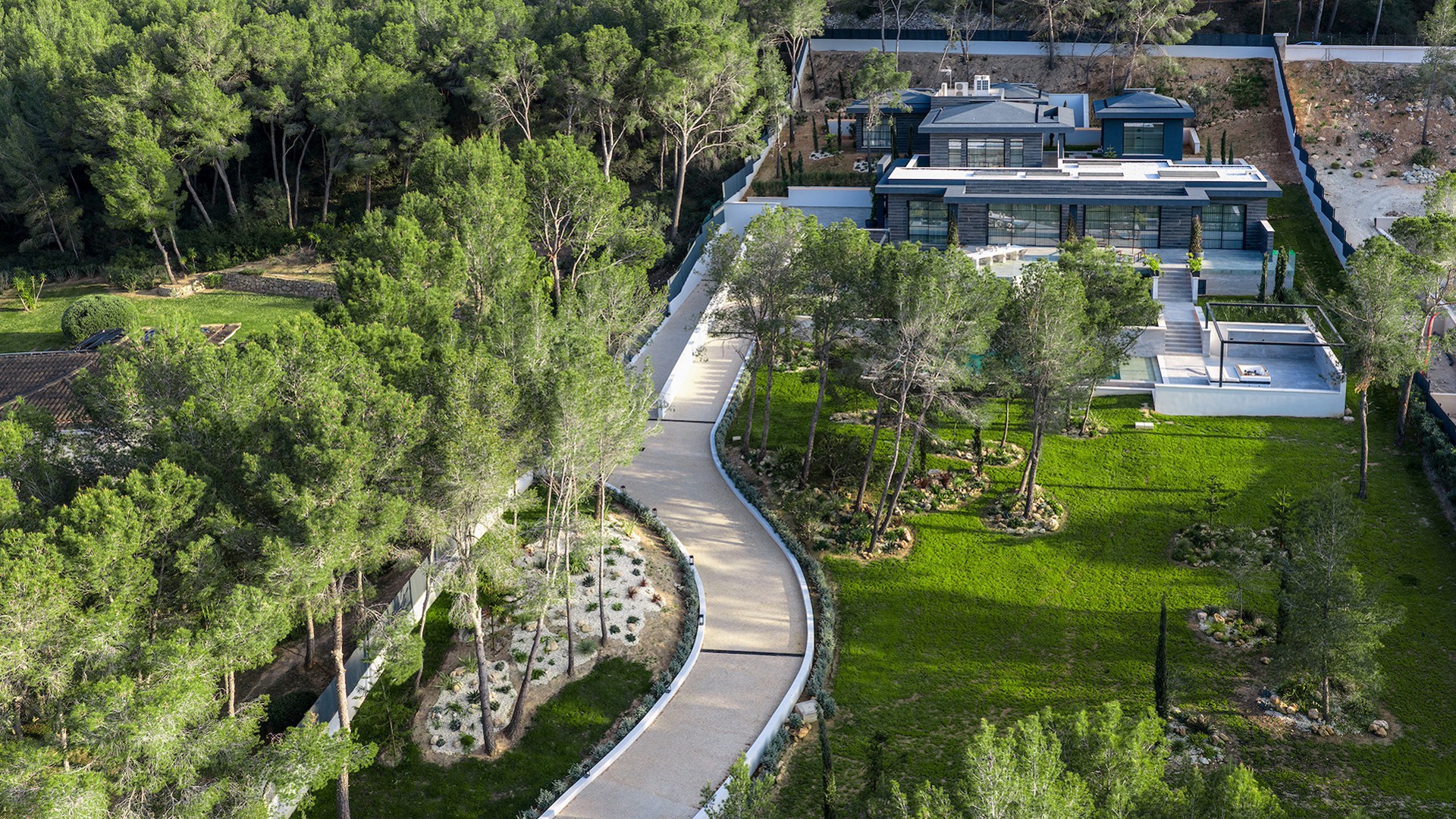 Aerial view of a modern villa with a flat roof, surrounded by gardens, pine trees, and a winding driveway.