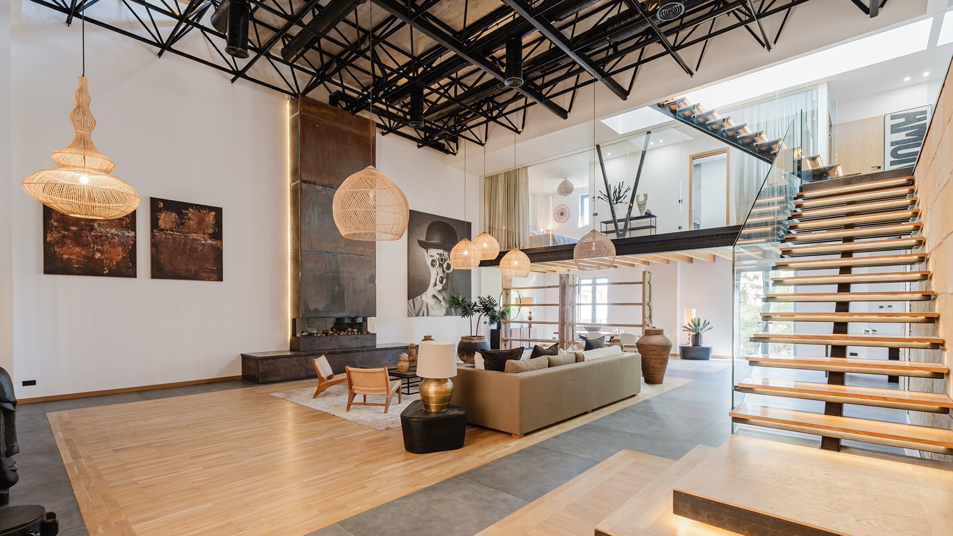A spacious apartment interior with high ceilings, exposed metal roof trusses, a floating staircase, and neutral-toned furniture.