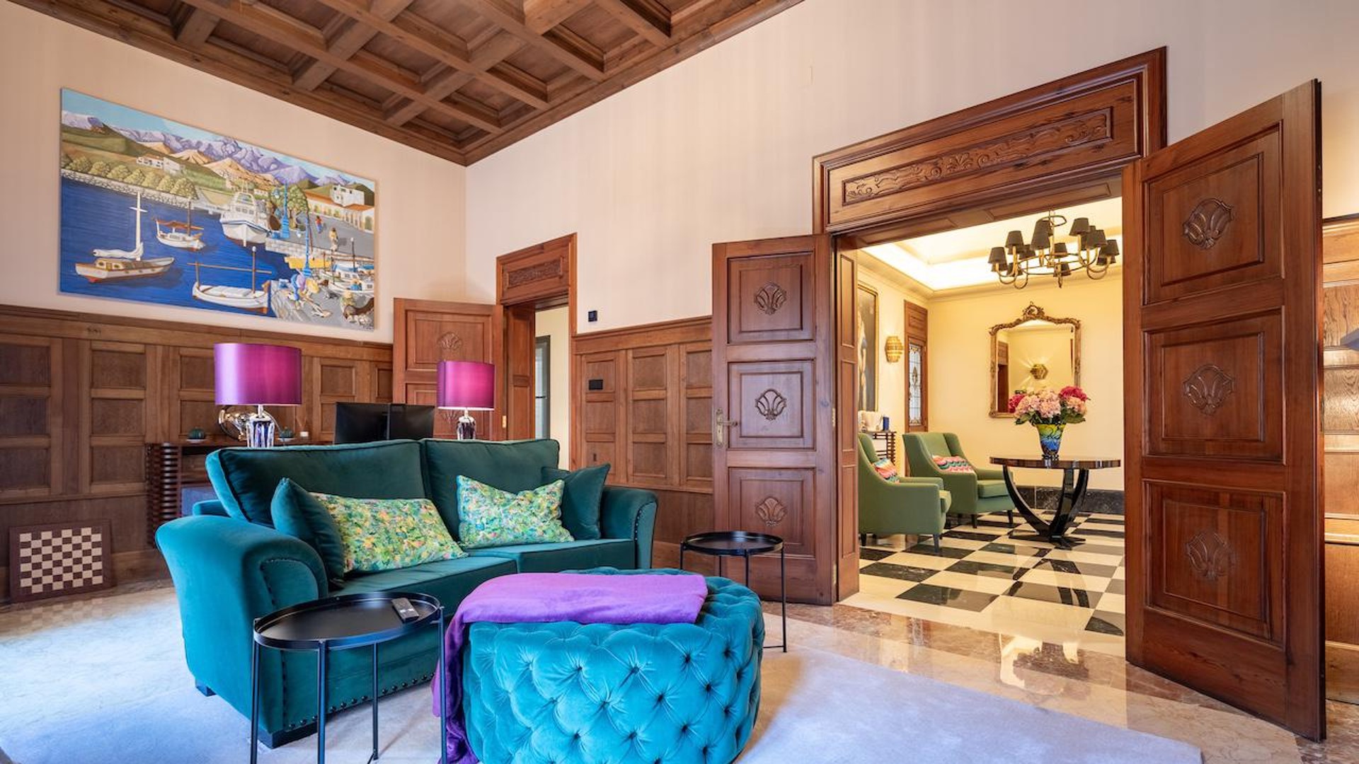 A formal living room with dark wood wall paneling, a coffered ceiling, a velvet teal sofa, and an open doorway leading to a tiled room.