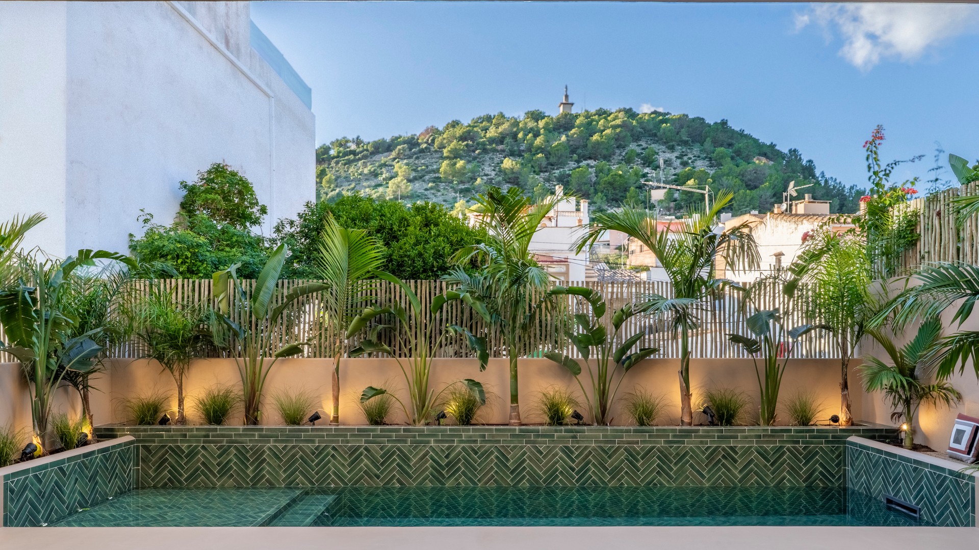 A rectangular pool with green chevron tiling and tropical plants, set against a backdrop of the Na Burguesa hills.