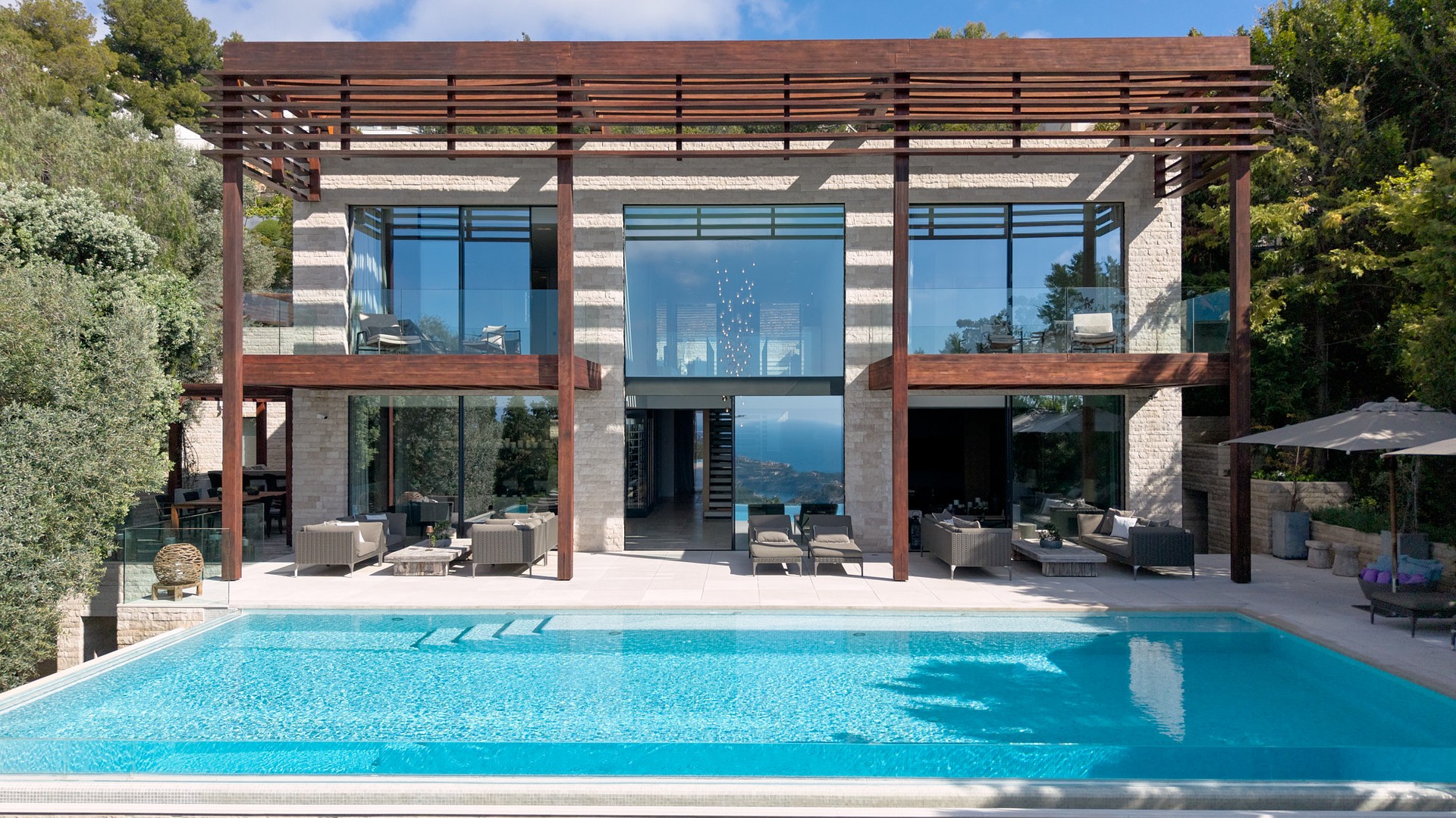 Exterior of a contemporary villa featuring an infinity pool, natural stone facade, floor-to-ceiling glass, and timber pergola.