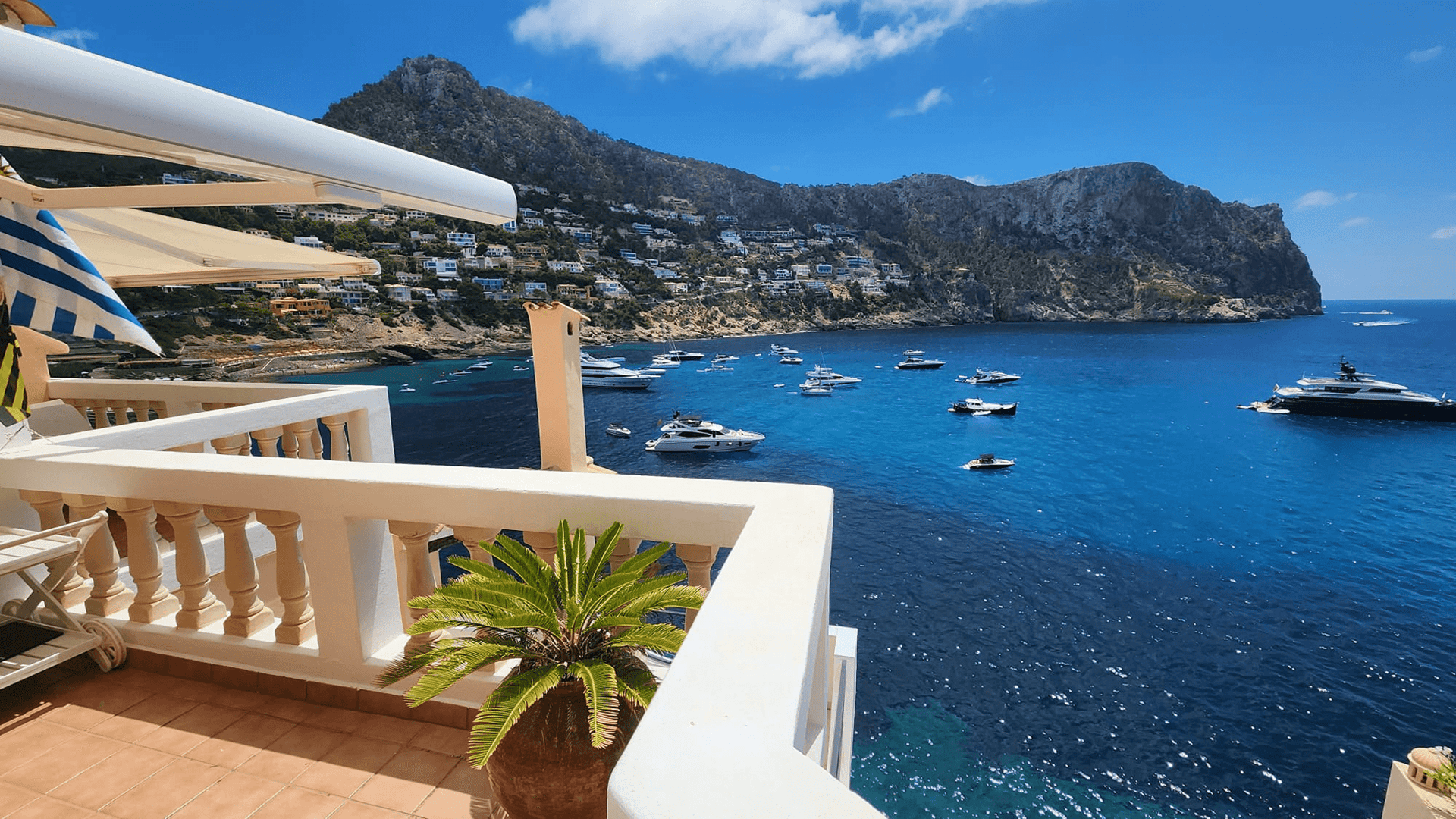 A wide-angle shot of a terrace with white balustrades overlooking the blue waters of Cala Llamp, featuring mountains and anchored boats.