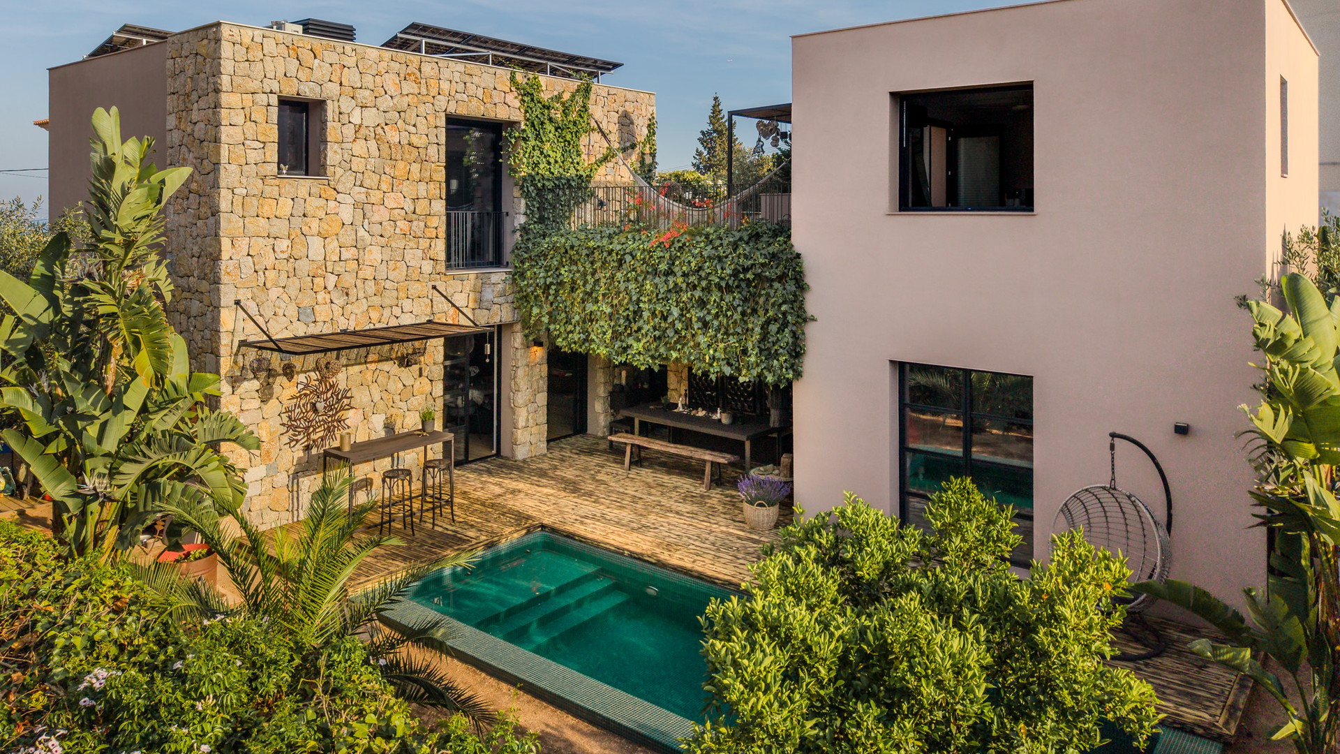 Exterior view of a multi-story villa in Genova featuring a natural stone wall section, smooth pink stucco, a swimming pool, and lush surrounding vegetation.