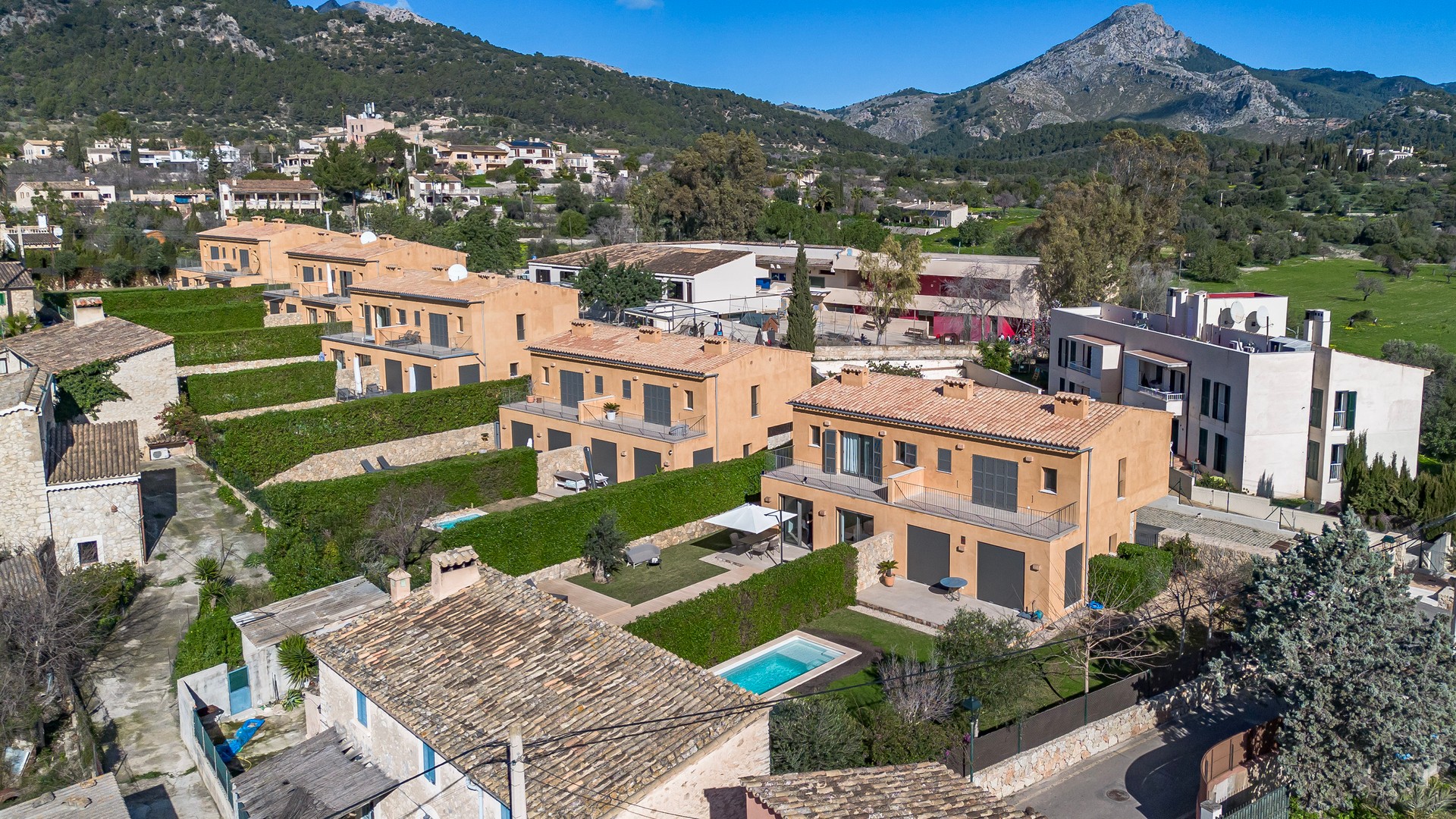 Aerial drone view showing a row of terraced townhouses with ochre facades, landscaped gardens, and a pool in Es Capdella.