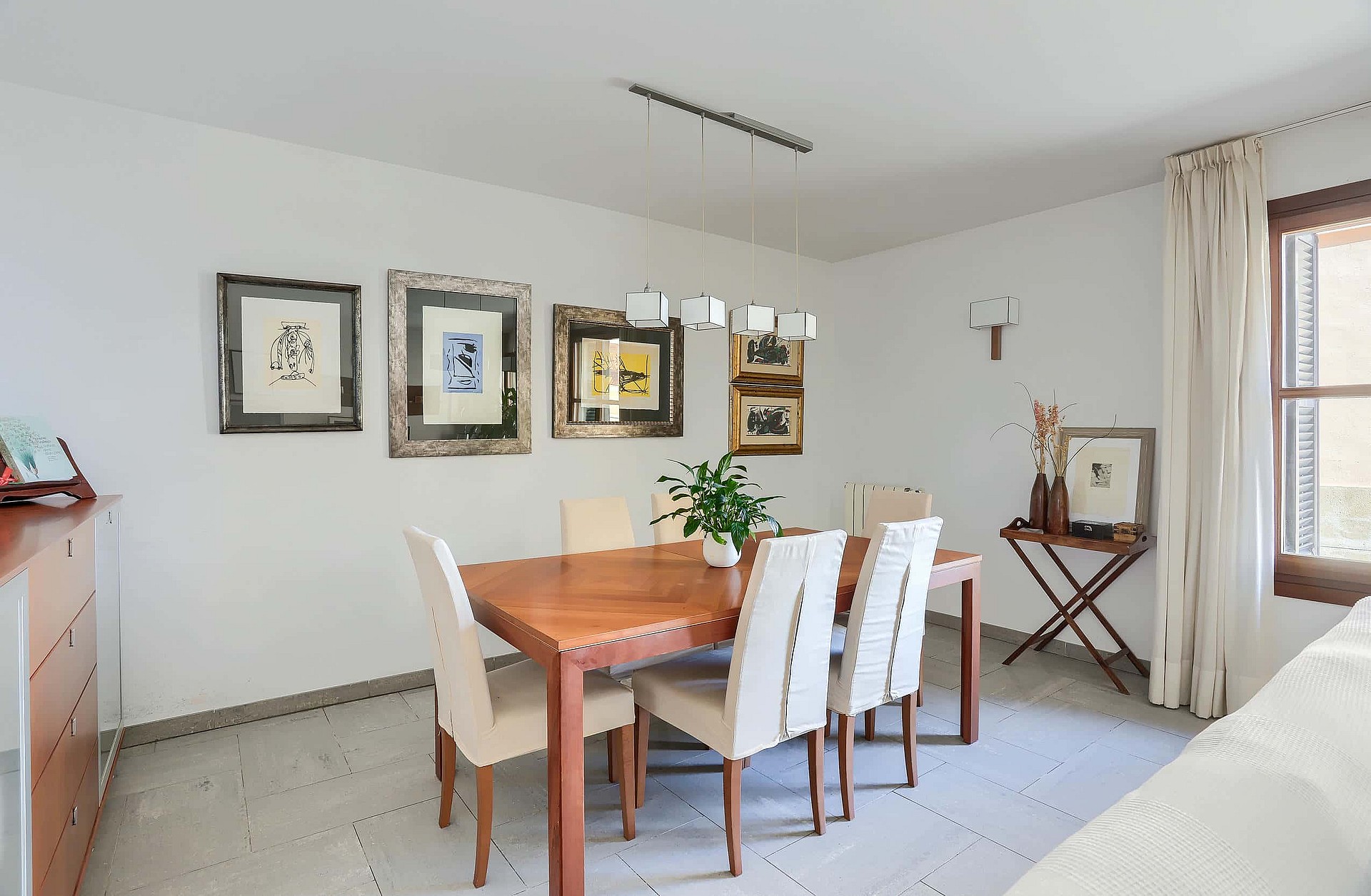 A dining room featuring a rectangular wooden table with six cream-upholstered chairs, white walls decorated with framed artwork, and a hanging pendant light.