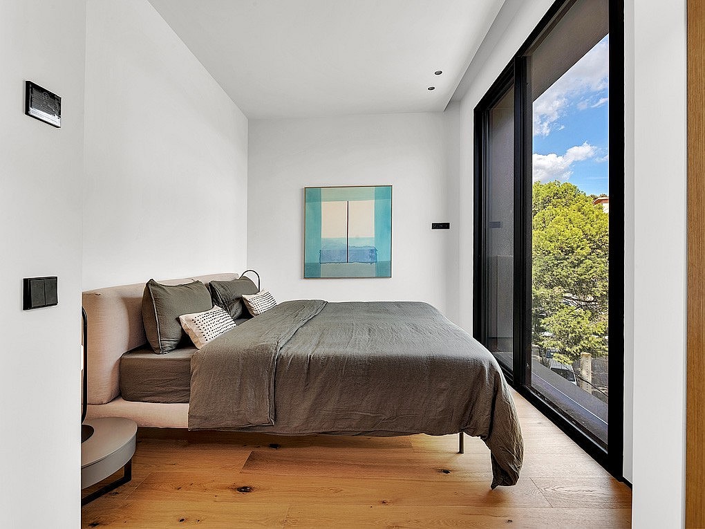Modern bedroom with oak wood flooring, gray bedding, and large windows overlooking green trees under a blue sky.
